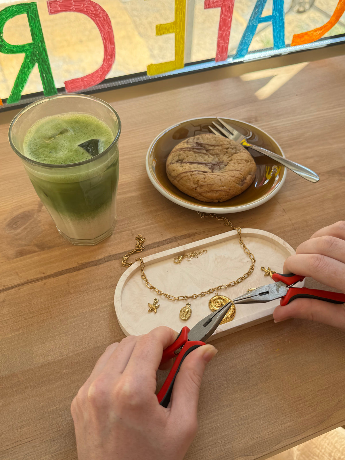 Bracelet à charms en cours de création, à côté d’un matcha et d’une viennoiserie, chez Mamie Jo à Nice